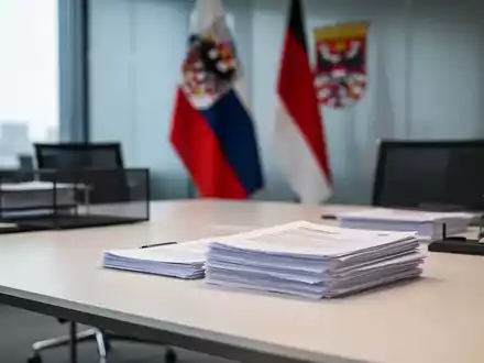Ein aufgeräumter Schreibtisch mit Dokumenten in einem modernen Regierungsbüro vor einer unscharfen Thüringer Flagge. - Foto: über boerse-global.de
