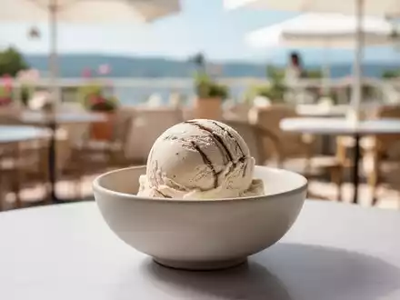 Eine einzelne Kugel Eiscreme in einer minimalistischen Schale vor einem unscharfen Hintergrund einer sommerlichen Terrasse. - Foto: über boerse-global.de