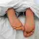Die nackten Füße und Pyjamahose einer Frau sind unter einer Bettdecke zu sehen. - Bild: Elisa Schu/dpa