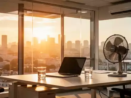 Ein moderner, heller Büroarbeitsplatz mit Ventilator und Wasserglas vor einem Fenster, durch das sommerliche Hitze schimmert. - Foto: über boerse-global.de