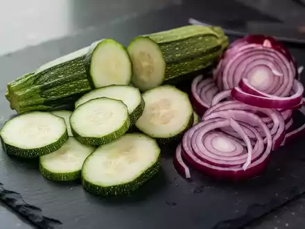 Frische Zucchini und rote Zwiebeln auf einer dunklen Schieferplatte, professionell in Szene gesetzt. - Foto: über boerse-global.de