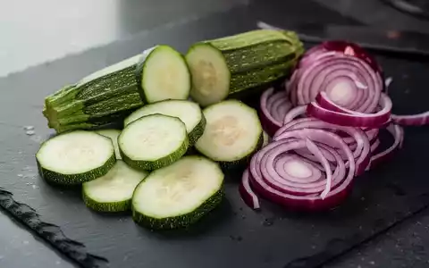 Frische Zucchini und rote Zwiebeln auf einer dunklen Schieferplatte, professionell in Szene gesetzt. - Foto: über boerse-global.de