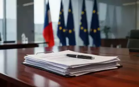 Dokumente auf einem Schreibtisch in einem Regierungsbüro mit EU-Flaggen im Hintergrund als Symbol für rechtliche Änderungen. - Foto: über boerse-global.de