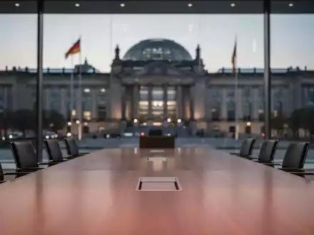 Ein moderner Konferenztisch vor dem unscharfen Reichstagsgebäude, symbolisch für politische Verhandlungen. - Foto: über boerse-global.de