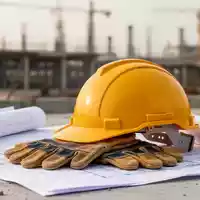 Ein gelber Arbeitsschutzhelm und Schutzhandschuhe auf Bauplänen vor einem unscharfen Hintergrund einer Baustelle. - Foto: über boerse-global.de