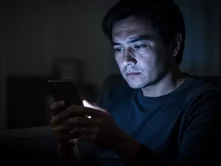 Eine Person blickt in einem abgedunkelten Raum auf ein leuchtendes Smartphone, das ihr Gesicht in den Fokus rückt. - Foto: über boerse-global.de