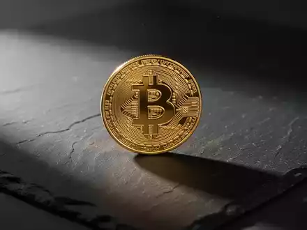 Eine einzelne goldene Bitcoin-Münze auf einer dunklen Schieferplatte bei stimmungsvoller Beleuchtung. - Foto: über boerse-global.de