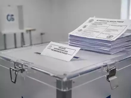 Nahaufnahme einer Wahlurne in einem hellen Wahllokal mit einem Stapel Stimmzettel im Hintergrund. - Foto: über boerse-global.de