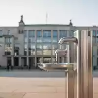 Moderne Trinkwasserstelle vor einem unscharfen Berliner Regierungsgebäude als Symbol für staatlichen Hitzeschutz. - Foto: über boerse-global.de