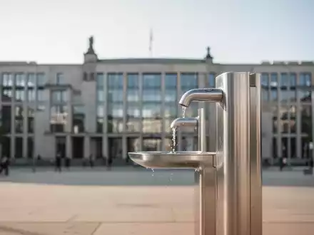 Moderne Trinkwasserstelle vor einem unscharfen Berliner Regierungsgebäude als Symbol für staatlichen Hitzeschutz. - Foto: über boerse-global.de