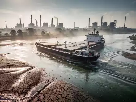 Ein fast ausgetrockneter Rhein mit einem schwer beladenen Binnenschiff vor einer industriellen Chemieanlage. - Foto: über boerse-global.de