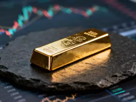 Ein glänzender Goldbarren auf einem dunklen Schieferuntergrund vor einem verschwommenen Finanzchart. - Foto: über boerse-global.de