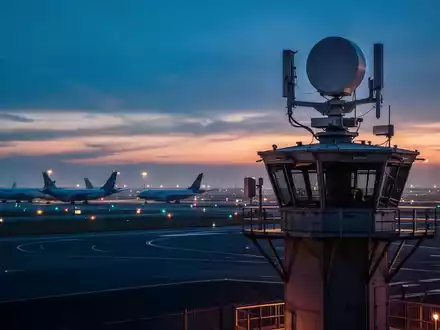 Ein Radarsystem auf einem Flughafentower bei Dämmerung mit Frachtflugzeugen im Hintergrund. - Foto: über boerse-global.de