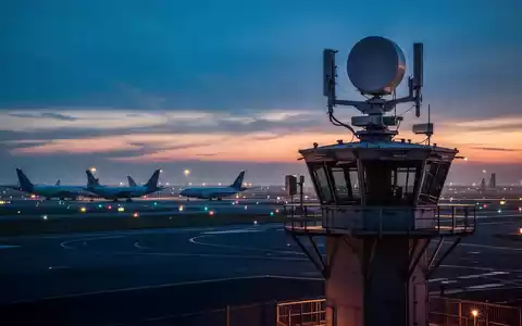 Ein Radarsystem auf einem Flughafentower bei Dämmerung mit Frachtflugzeugen im Hintergrund. - Foto: über boerse-global.de