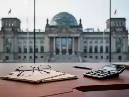 Ein Schreibtisch mit Brille und Taschenrechner vor einem unscharfen Hintergrund des Reichstagsgebäudes. - Foto: über boerse-global.de