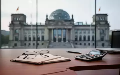 Ein Schreibtisch mit Brille und Taschenrechner vor einem unscharfen Hintergrund des Reichstagsgebäudes. - Foto: über boerse-global.de