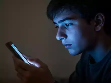 Ein junger Mensch blickt in einem abgedunkelten Raum auf ein leuchtendes Smartphone, das sein Gesicht in kaltes Licht taucht. - Foto: über boerse-global.de