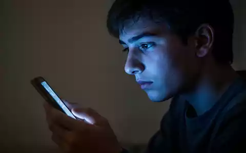 Ein junger Mensch blickt in einem abgedunkelten Raum auf ein leuchtendes Smartphone, das sein Gesicht in kaltes Licht taucht. - Foto: über boerse-global.de