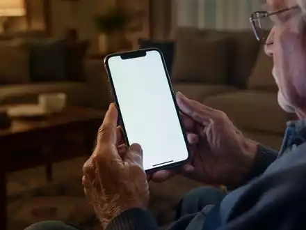 Ein älterer Mann hält ein Smartphone in den Händen, während er in einem Wohnzimmer sitzt. - Foto: über boerse-global.de