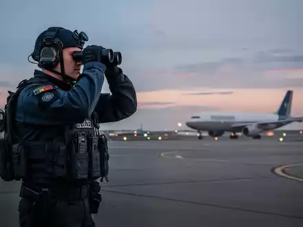 Ein spezialisierter Bundespolizist in taktischer Ausrüstung überwacht bei Dämmerung das Gelände eines Flughafens. - Foto: über boerse-global.de