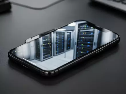 Ein Smartphone auf einem dunklen Schreibtisch vor dem unscharfen Hintergrund eines Serverraums. - Foto: über boerse-global.de