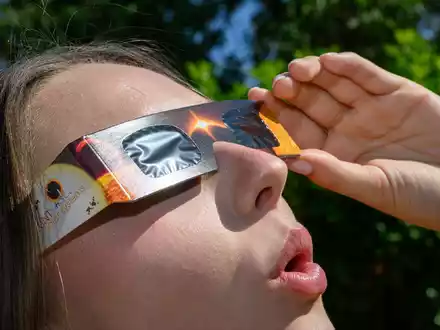 Eine junge Frau schaut durch eine Sonnenfinsternisbrille und betrachtet die Mittagssonne. - Foto: Patrick Pleul/dpa