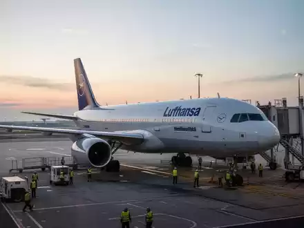Ein Lufthansa-Flugzeug steht bei Dämmerung auf dem Rollfeld eines Flughafens, während Bodenpersonal das Flugzeug vorbereitet. - Foto: über boerse-global.de