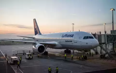 Ein Lufthansa-Flugzeug steht bei Dämmerung auf dem Rollfeld eines Flughafens, während Bodenpersonal das Flugzeug vorbereitet. - Foto: über boerse-global.de