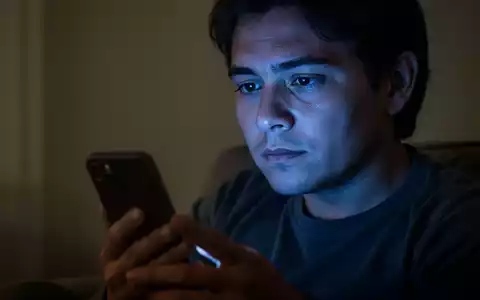 Eine Person sitzt in einem abgedunkelten Raum und schaut nachdenklich auf ihr Smartphone, dessen Licht ihr Gesicht sanft beleuchtet. - Foto: über boerse-global.de