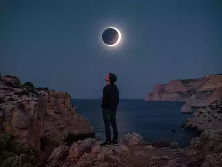 Eine Person betrachtet in kontemplativer Haltung eine totale Sonnenfinsternis ĂŒber einer felsigen KĂŒstenlandschaft. - Foto: ĂŒber boerse-global.de