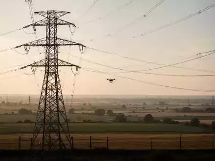 Ein großer Hochspannungsmast in einer weiten Landschaft bei Sonnenuntergang, in der Ferne ist schemenhaft eine Drohne zu sehen. - Foto: über boerse-global.de