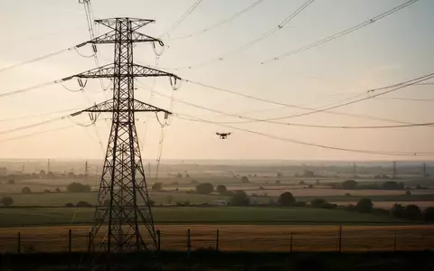 Ein großer Hochspannungsmast in einer weiten Landschaft bei Sonnenuntergang, in der Ferne ist schemenhaft eine Drohne zu sehen. - Foto: über boerse-global.de