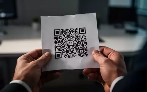 Nahaufnahme von Händen, die ein Blatt Papier mit einem QR-Code halten, in einer modernen Büroumgebung. - Foto: über boerse-global.de