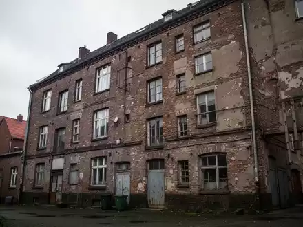 Ein sanierungsbedürftiges Wohngebäude in einem Industriegebiet unter einem bewölkten Himmel. - Foto: über boerse-global.de