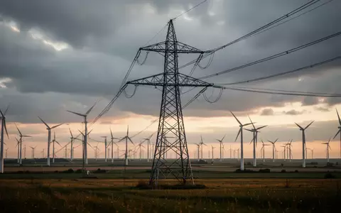 Ein Hochspannungsmast steht in einer weiten Landschaft mit Windkraftanlagen unter einem bewölkten Himmel. - Foto: über boerse-global.de