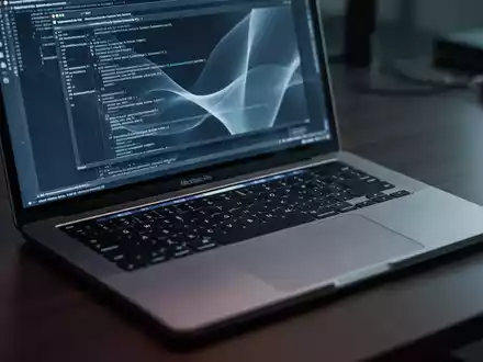 Ein MacBook Pro auf einem dunklen Schreibtisch, dessen Bildschirm eine abstrakte Terminal-Oberfläche zeigt, die Cybersicherheit symbolisiert. - Foto: über boerse-global.de