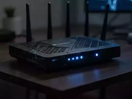 Ein moderner Internet-Router mit leuchtenden Status-LEDs auf einem dunklen Schreibtisch in einer schummrigen Umgebung. - Foto: über boerse-global.de