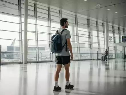 Eine minimalistisch ausgerüstete Reisende mit kleinem Rucksack in einem modernen, lichtdurchfluteten Flughafen-Terminal. - Foto: über boerse-global.de