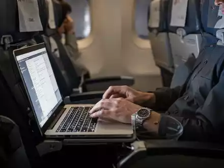 Ein Passagier sitzt in einem Flugzeug und arbeitet an einem Laptop, während das Licht des Bildschirms das Gesicht beleuchtet. - Foto: über boerse-global.de