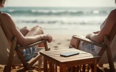 Ein Smartphone liegt mit dem Display nach unten auf einem Holztisch an einem sonnigen Strand, während der Besitzer entspannt. - Foto: über boerse-global.de