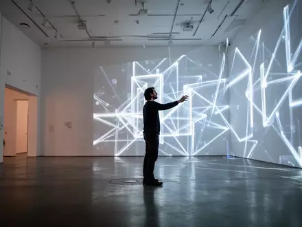 Eine Person interagiert in einer minimalistischen Berliner Galerie mit einer großformatigen, leuchtenden digitalen Kunstinstallation. - Foto: über boerse-global.de