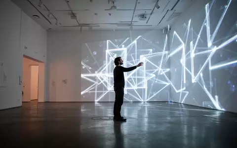 Eine Person interagiert in einer minimalistischen Berliner Galerie mit einer großformatigen, leuchtenden digitalen Kunstinstallation. - Foto: über boerse-global.de