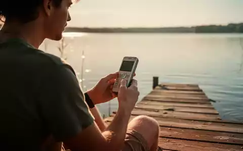 Eine Person sitzt am Sommertag an einem See und hält ein schlichtes Tastenhandy in der Hand. - Foto: über boerse-global.de