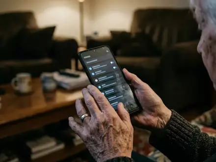 Nahaufnahme der Hände eines Senioren, der ein Smartphone in einem abgedunkelten Raum hält, während er auf den Bildschirm blickt. - Foto: über boerse-global.de