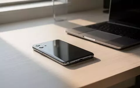 Ein modernes Smartphone liegt auf einem hellen Holztisch neben einem Laptop in einem aufgeräumten, lichtdurchfluteten Büro. - Foto: über boerse-global.de