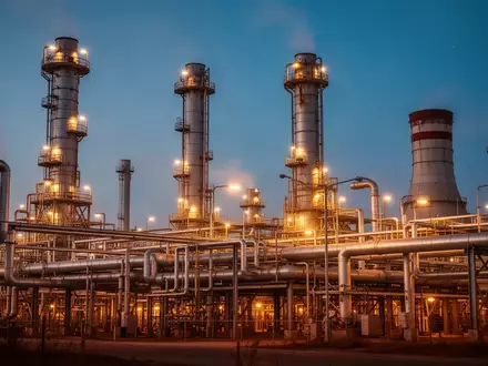 Industrielle Ölraffinerie bei Dämmerung mit warm beleuchteten Rohrsystemen und Kühltürmen. - Foto: über boerse-global.de
