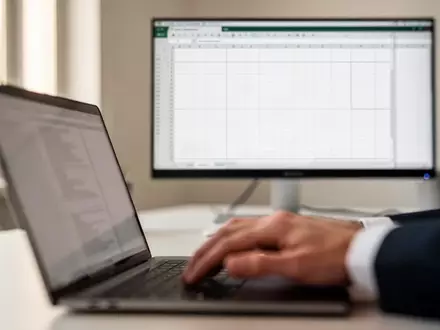 Ein professioneller Arbeitsplatz mit einem Laptop und einem Monitor, auf dem ein leeres Tabellenkalkulationsgitter zu sehen ist. - Foto: über boerse-global.de