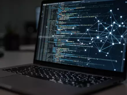 Ein Laptop-Bildschirm mit verschwommenem Programmiercode und Netzwerkverbindungen in einem abgedunkelten Büro. - Foto: über boerse-global.de