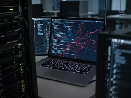 Ein Laptop-Bildschirm zeigt abstrakte Codezeilen in einem dunklen Büro, was auf IT-Sicherheitsrisiken und Cyberangriffe hindeutet. - Foto: über boerse-global.de