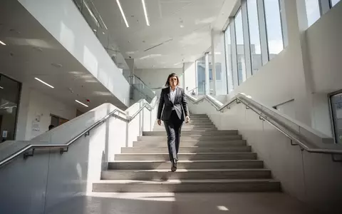 Eine Person steigt in einem hellen, modernen Treppenhaus eine Treppe hinauf, Tageslicht durchflutet das Gebäude. - Foto: über boerse-global.de